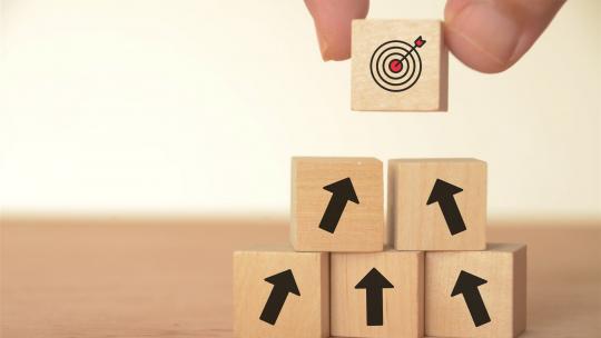 Holzklötze mit Pfeilsymbolen, die aufwärts zeigen, werden zu einer Pyramide gestapelt; oberster Block mit Zielscheibe symbolisiert strategische Zielerreichung im HR-Prozess