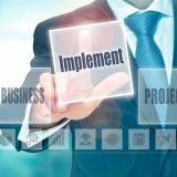 Business-Mann in Anzug tippt auf ein transparentes digitales Interface mit dem Begriff „Implement“ – symbolisiert unsere Umsetzung von Business-Projekten und strategischen Maßnahmen